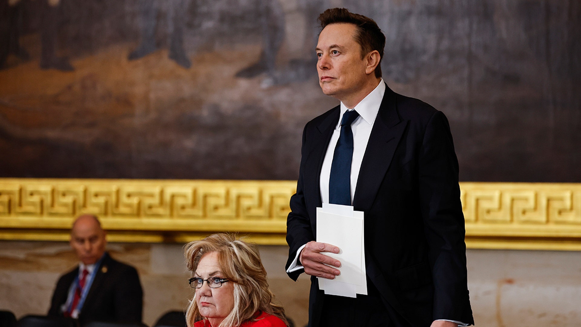Elon Musk in a dark suit with a paper to his chest stands in the Rotunda