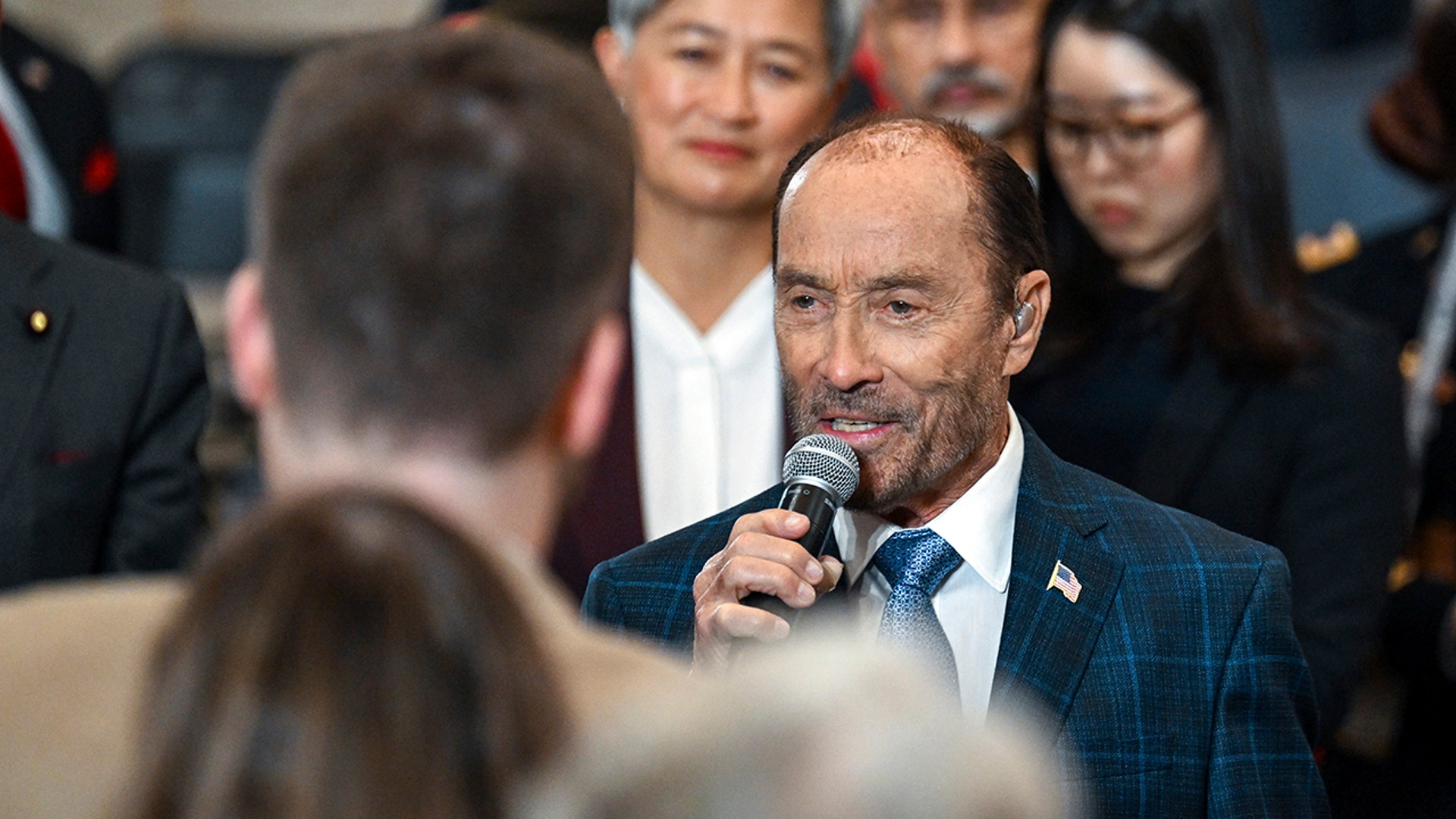 Lee Greenwood in a blue/turquoise plaid jacket sings into the microphone ahead of the inauguration
