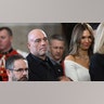 Joe Rogan in a black suit looks serious in the Rotunda as he stands for benediction with his wife Jessica