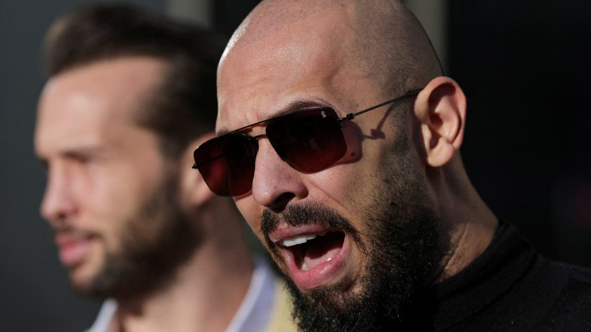 Andrew Tate speaking in a closeup image, he is wearing a black shirt and sunglasses, is bald and has a bushy goatee