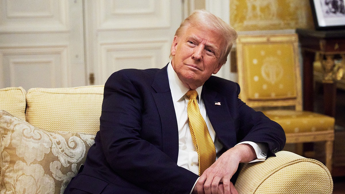 Trump wearing a dark blue suit and a yellow tie sitting on a yellow couch.