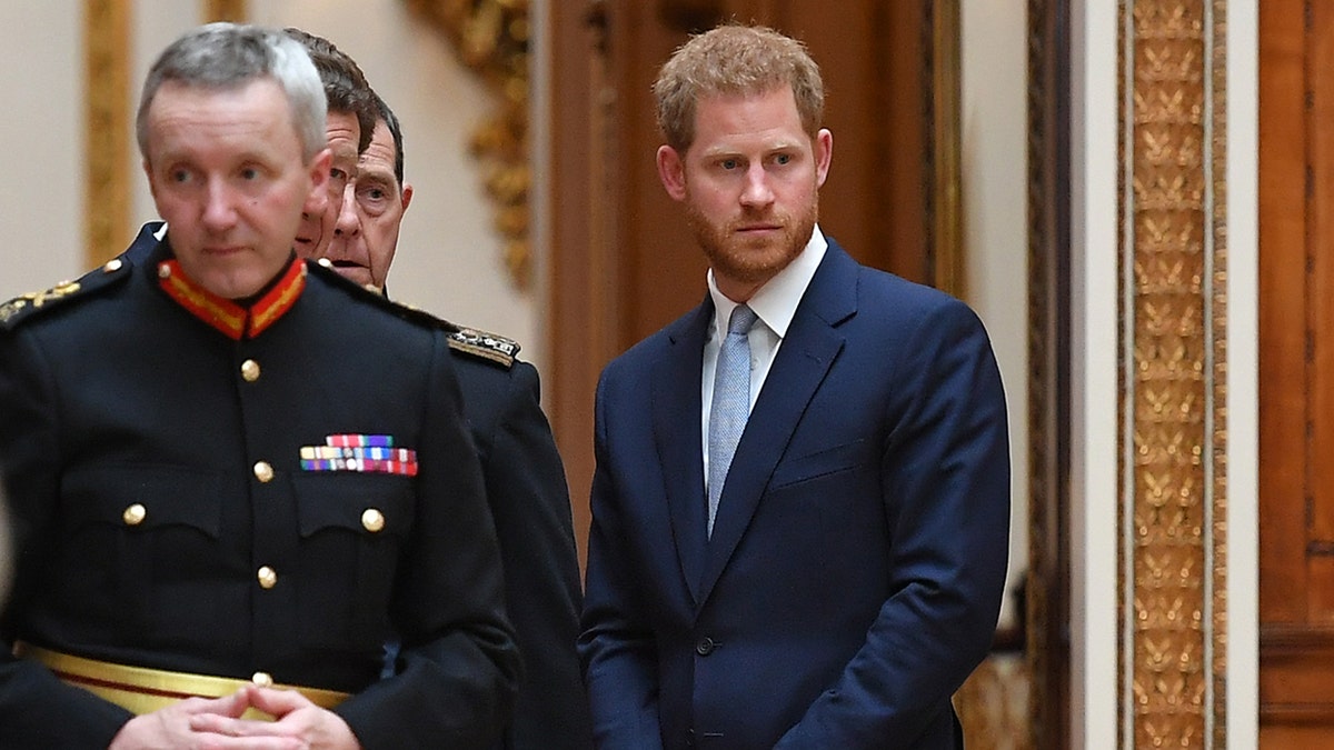 Prince Harry wearing a blue blazer and a white collared shirt with a light blue tie standing next to several men in uniform.