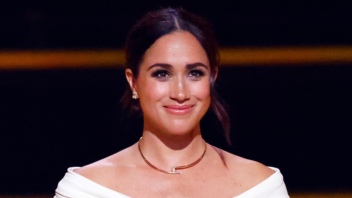 A close-up of Meghan Markle smiling with a gold necklace and a white top.