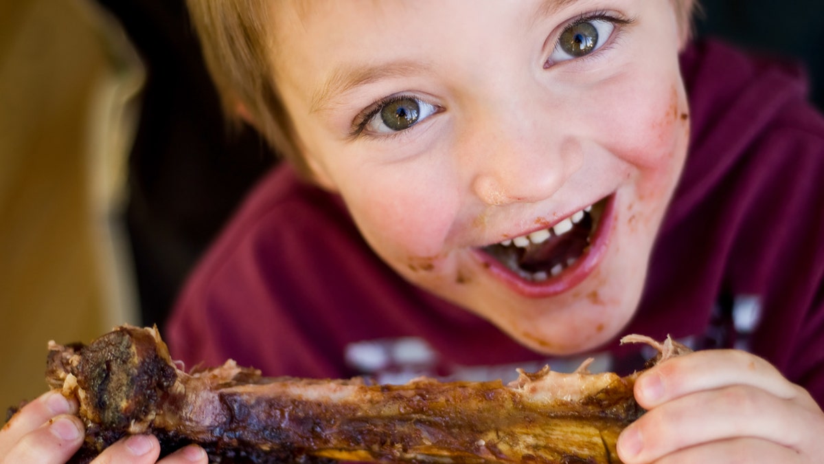 A boy with a messy face smiles as he holds a rib in his hand.