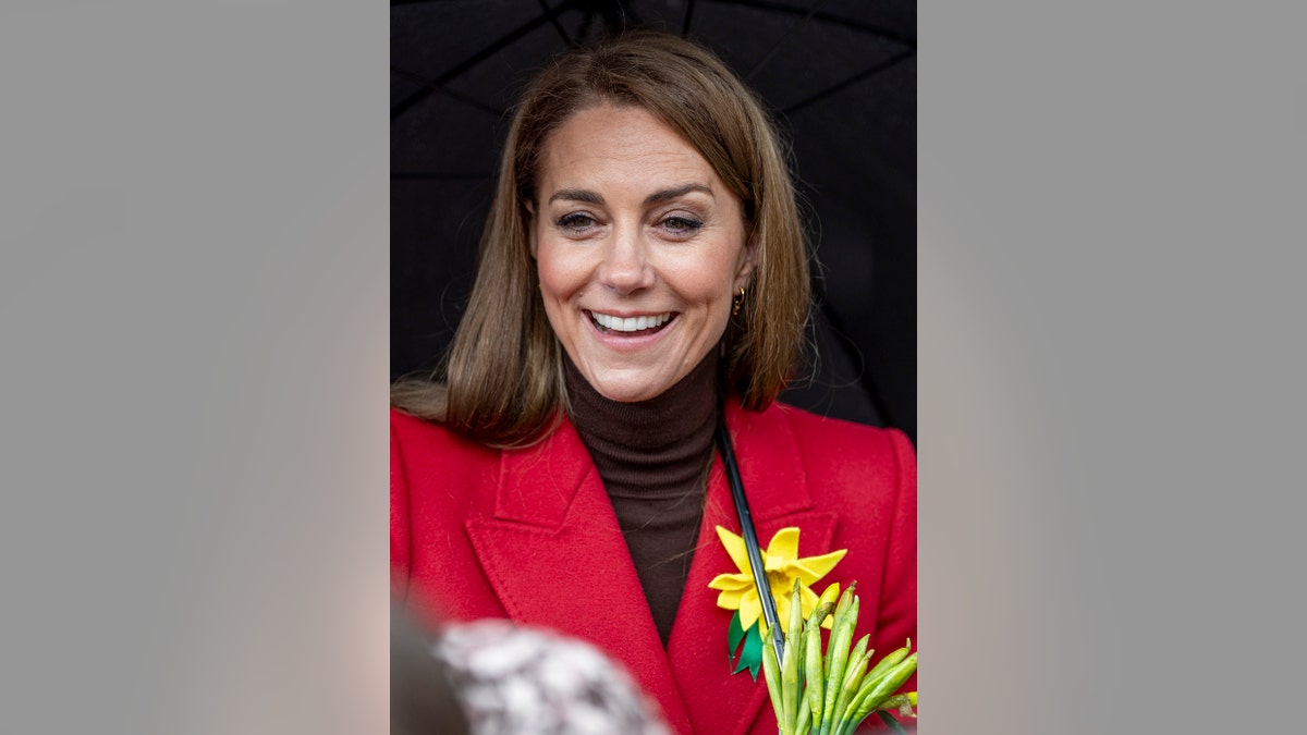 A close-up of Kate Middleton wearing a brown turtleneck and a red coat smiling with a daffodil