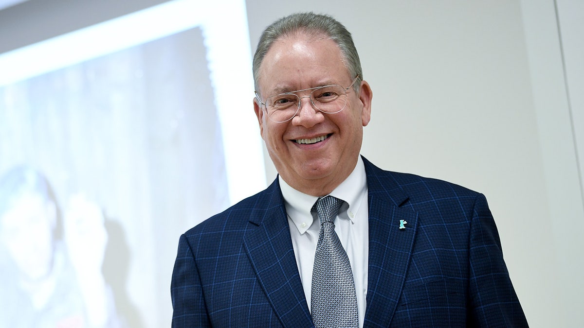 Chris Lewis wearing a blue blazer, a white shirt and a blue and silver tie as he smiles inside a white building.