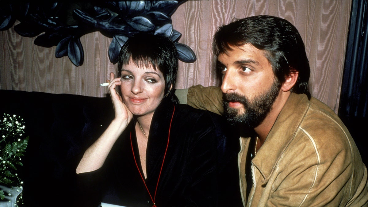 Liza Minnelli smiling wearing a black robe and holding a cigarette admiring Mark Gero wearing a gold blazer