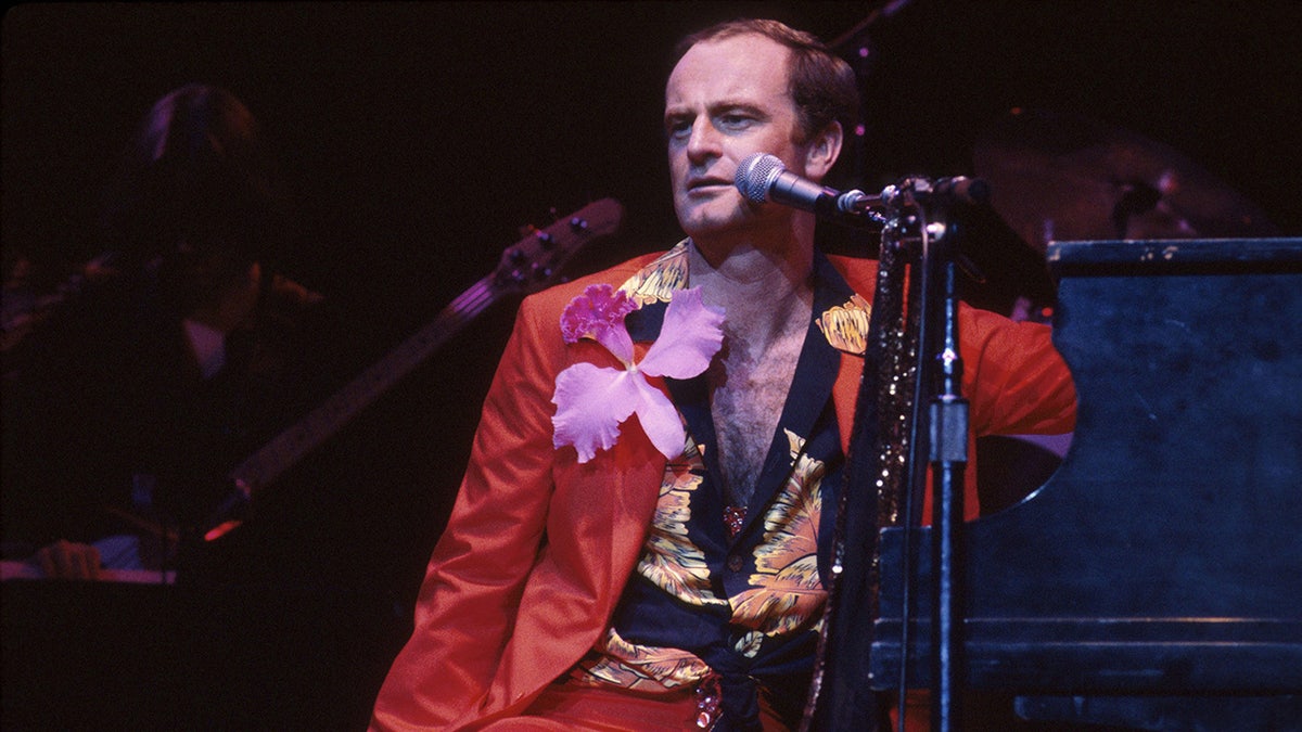 Peter Allen wearing a coral suit with a floral shirt and a purple orchid brooch on stage next to a mic.