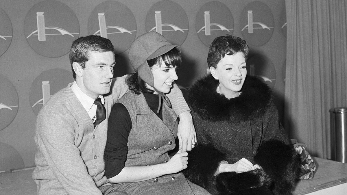 Peter Allen with his arm around Liza Minnelli as she sits next to her mother Judy Garland, who is wearing a fur coat and smiling.