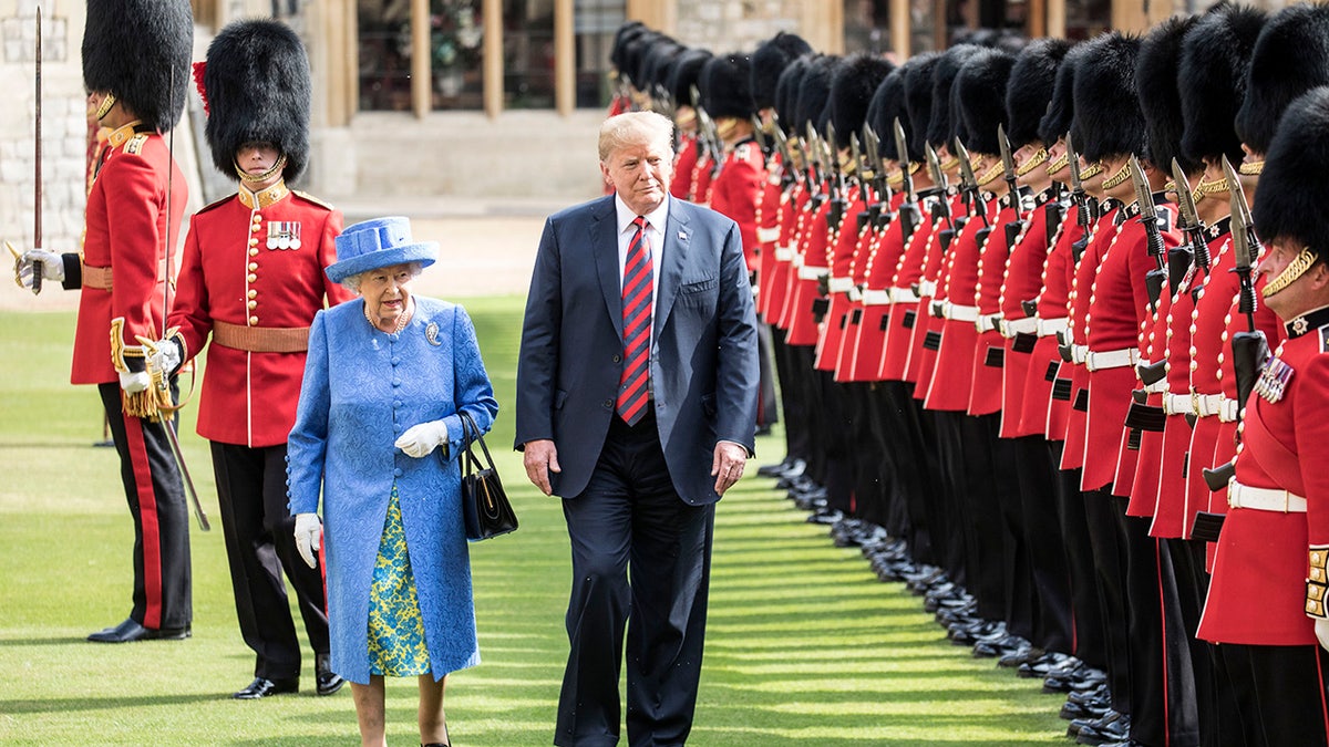 Queen Elizabeth in a blue coat and floral dress walking next to Trump in a suit and a red and blue tie
