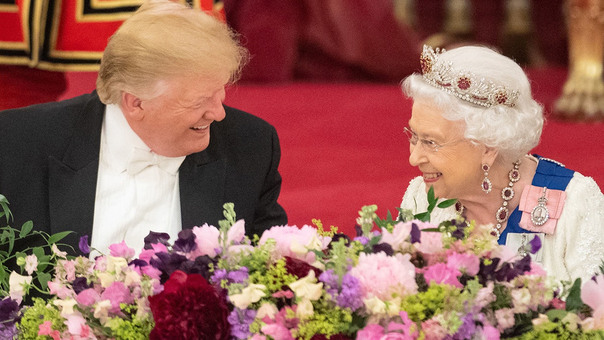 President Trump in a black blazer and white shirt sitting next to Queen Elizabeth in a white dress with a tiara.