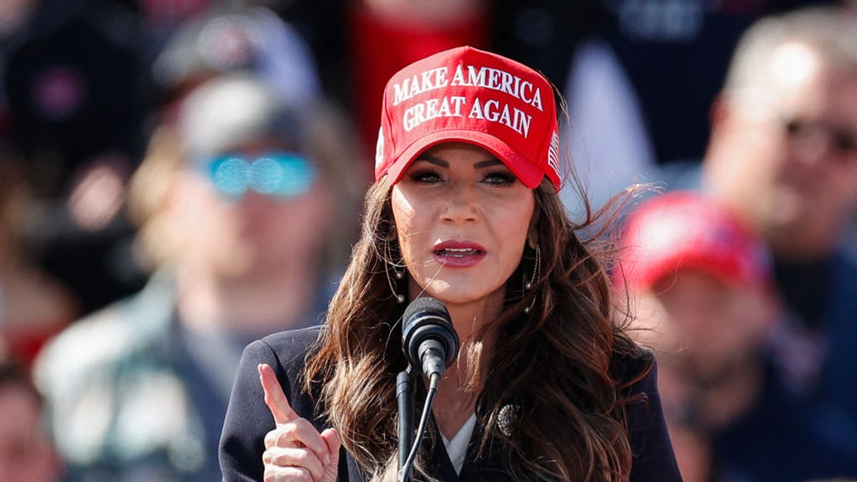 Noem speaking at a rally wearing a red MAGA hat.