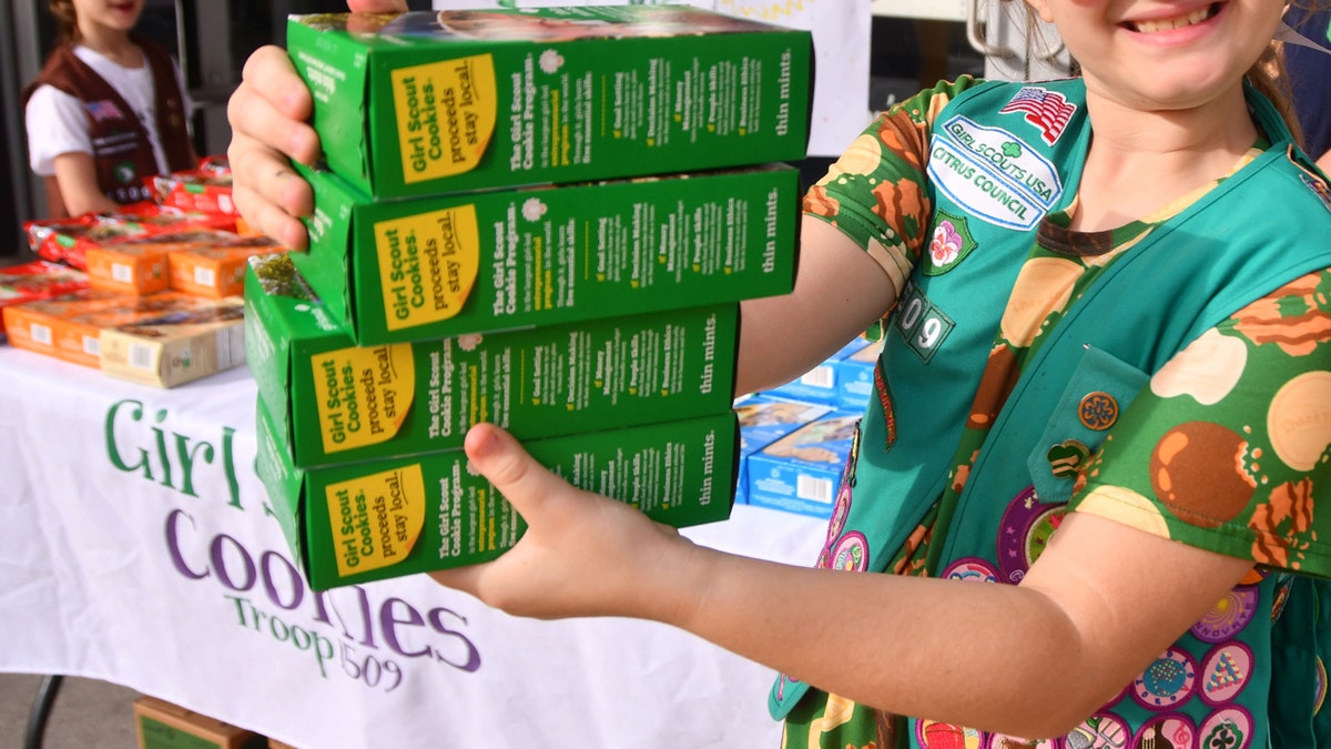 A Girl Scout holds four boxes of Thin Mints at a table where Girl Scout cookies are sold.