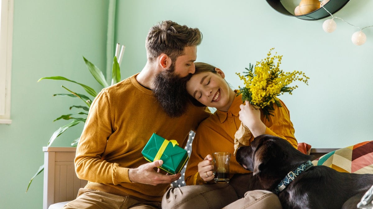 Portrait of affectionate young man embracing and kissing his loving girlfriend after giving her mimosa flowers and a gift box.