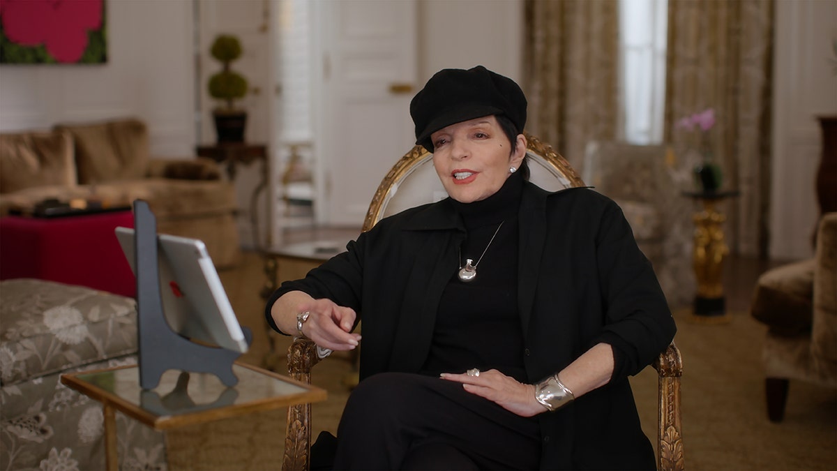 Liza Minnelli wearing all black with a matching hat and a silver necklace sitting down and smiling.