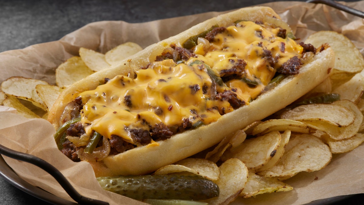 A Philadelphia cheesesteak sandwich is shown with potato chips and a pickle.