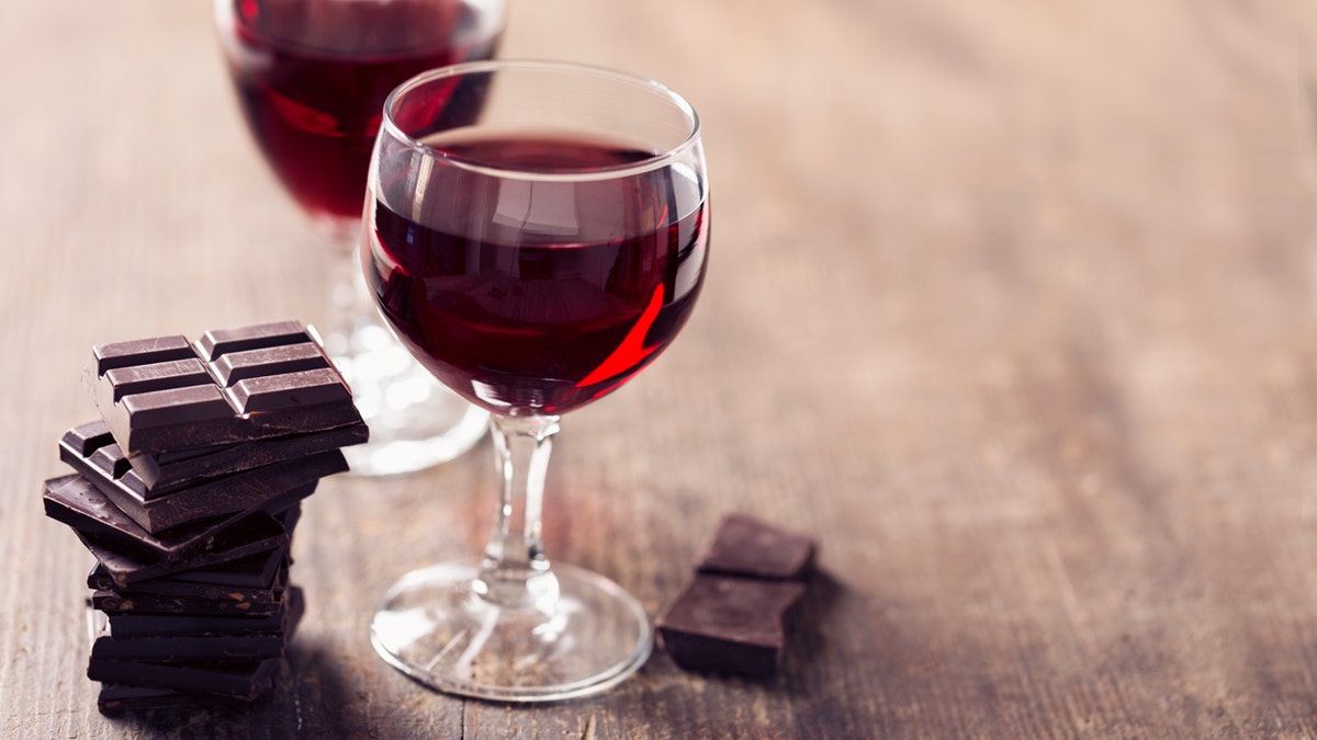 Two glasses of red wine are shown on a table next to pieces of chocolate.