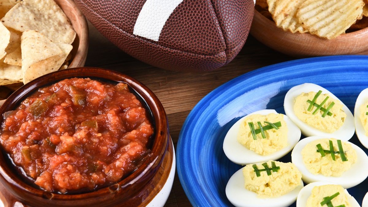 The stuffed eggs decorated to seem that the football is shown on a plate on a table next to football, french fries and sauce.