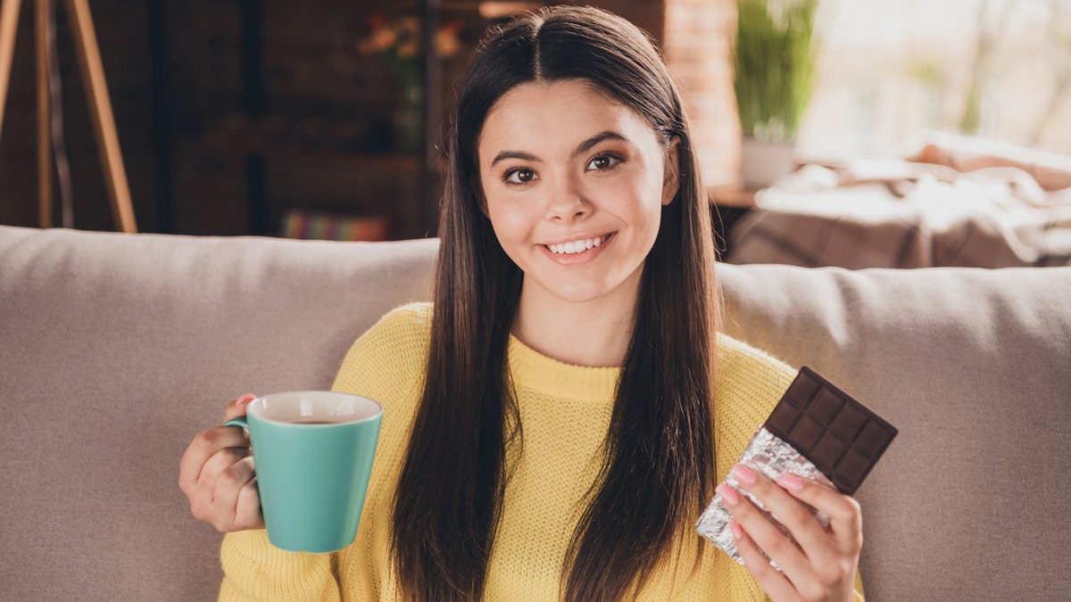 A woman smiles as she sits on a couch holding a cup of tea in one hand and a chocolate bar in the other.