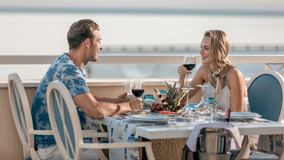 Two young couple drink red wine while eating outside the waterfront.