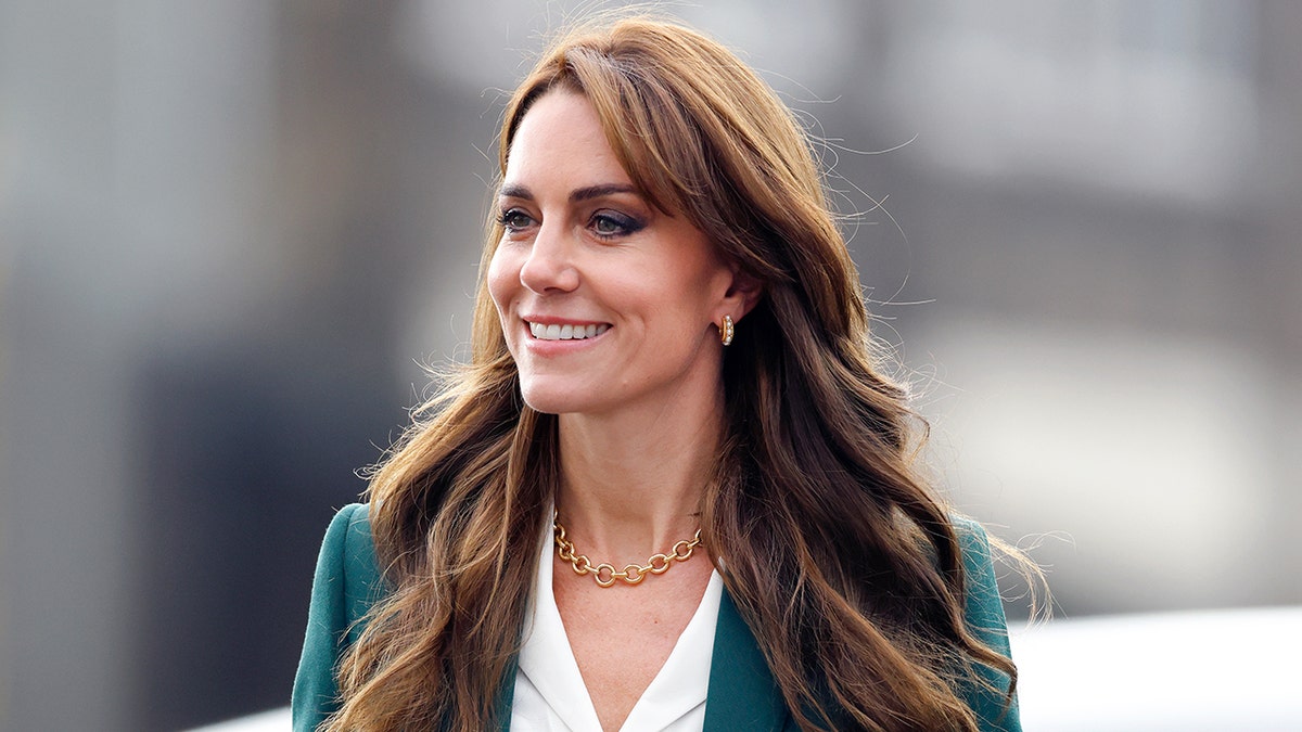 Kate Middleton looking to the side smiling in a green blazer and white shirt.