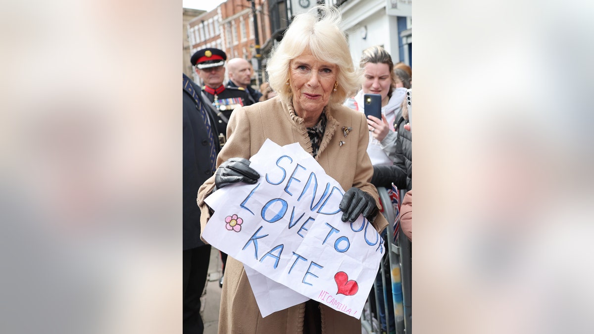 Queen Camilla holding a sign for Kate Middleton outdoors surrounded by people.