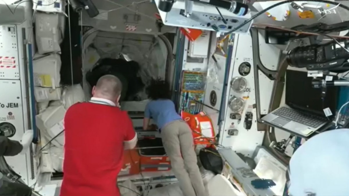 The astronaut uses an alien mask while crew members work inside the station