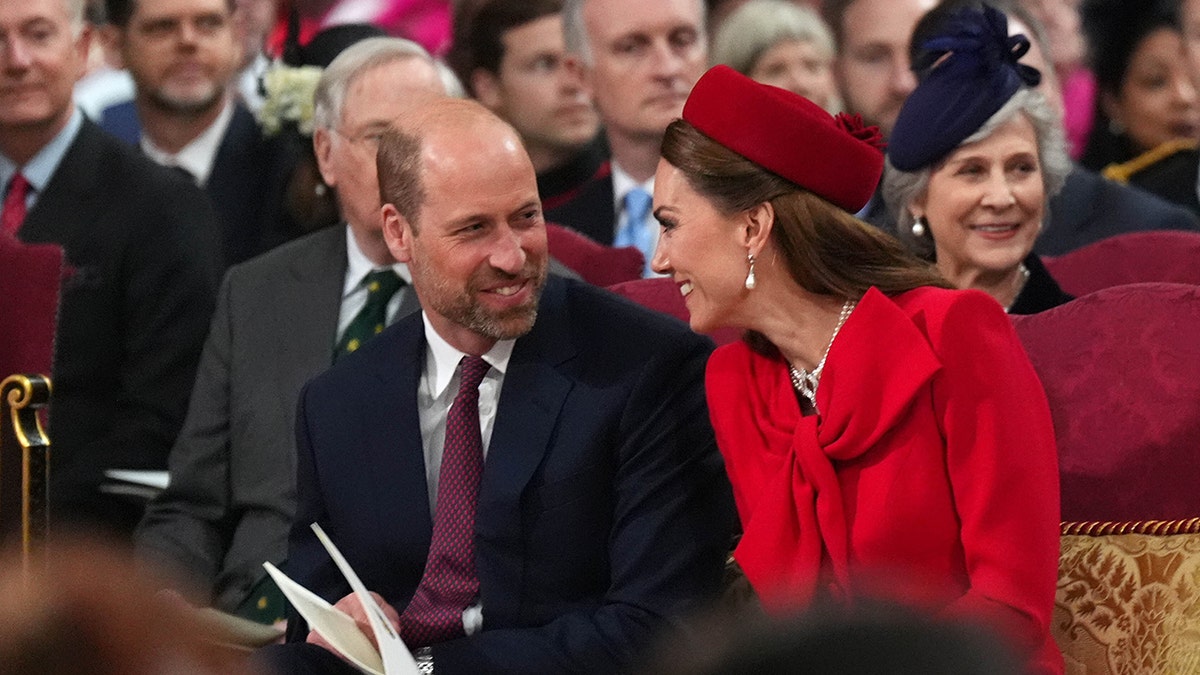 Kate Middleton wearing a red dress and a matching hat smiling next to a Prince William in a suit and purple tie.