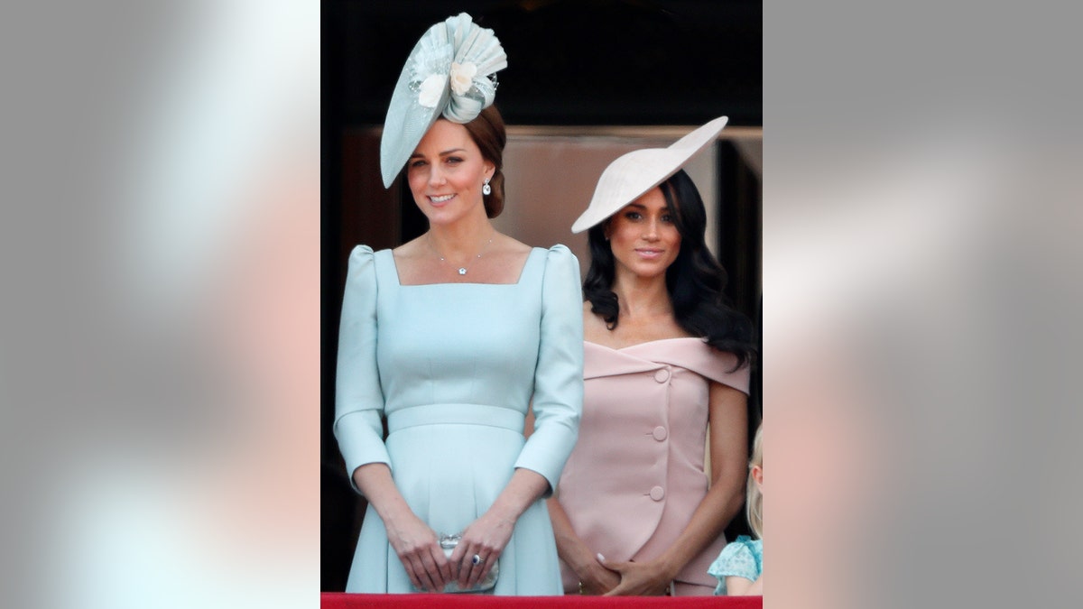 Kate Middleton wearing a light blue dress and a matching hat standing in front of Meghan Markle wearing a powder pink dress and a matching hat.