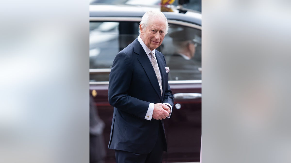 King Charles wearing a blue blazer and a white shirt with a tie standing next to a dark car.