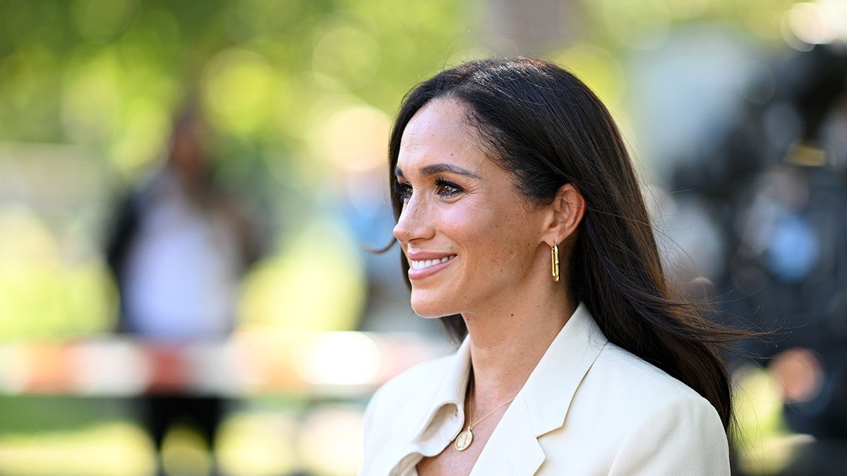 Meghan Markle smiled as she looked at the side of her face wearing an ivory blazer and gold jewelry.