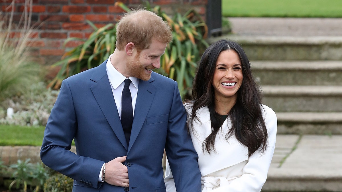 Prince Harry in a blue suit smiling outdoors with Meghan Markle wearing a white coat and a black dress.