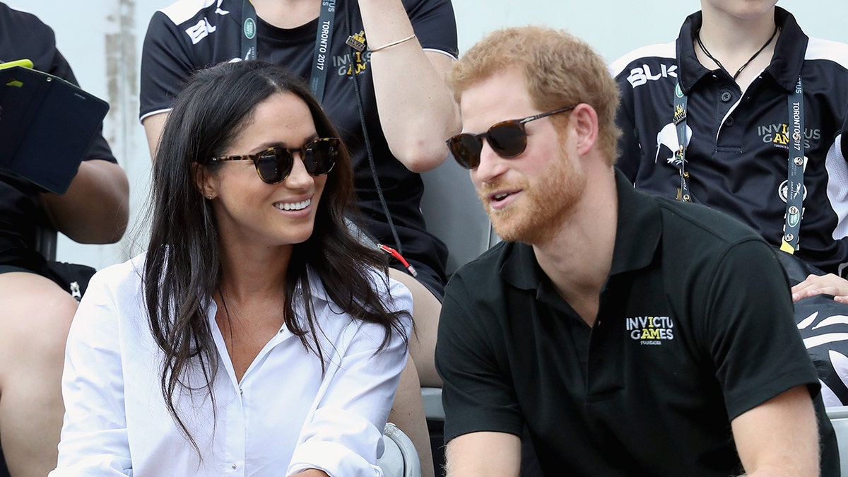 Meghan Markle wearing a white shirt and sunglasses sitting next to Prince Harry in a dark Invictus Games shirt and sunglasses.