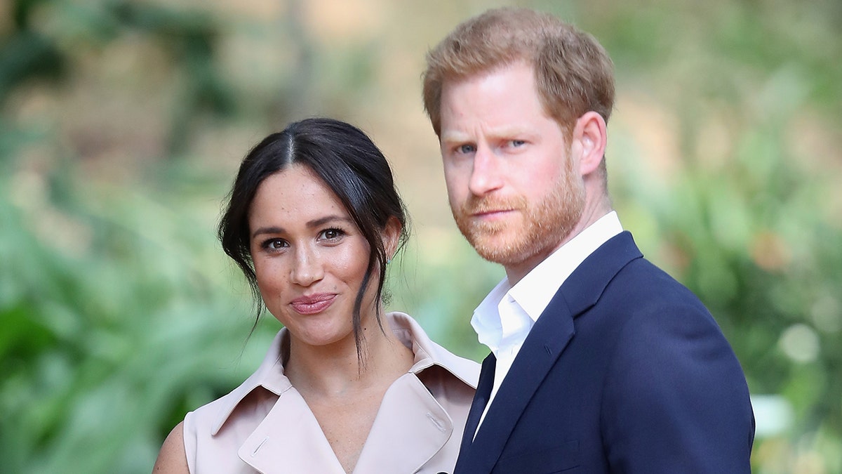 A close up of Meghan Markle wearing a beige dress next to Prince Harry in a blue suit and white shirt.