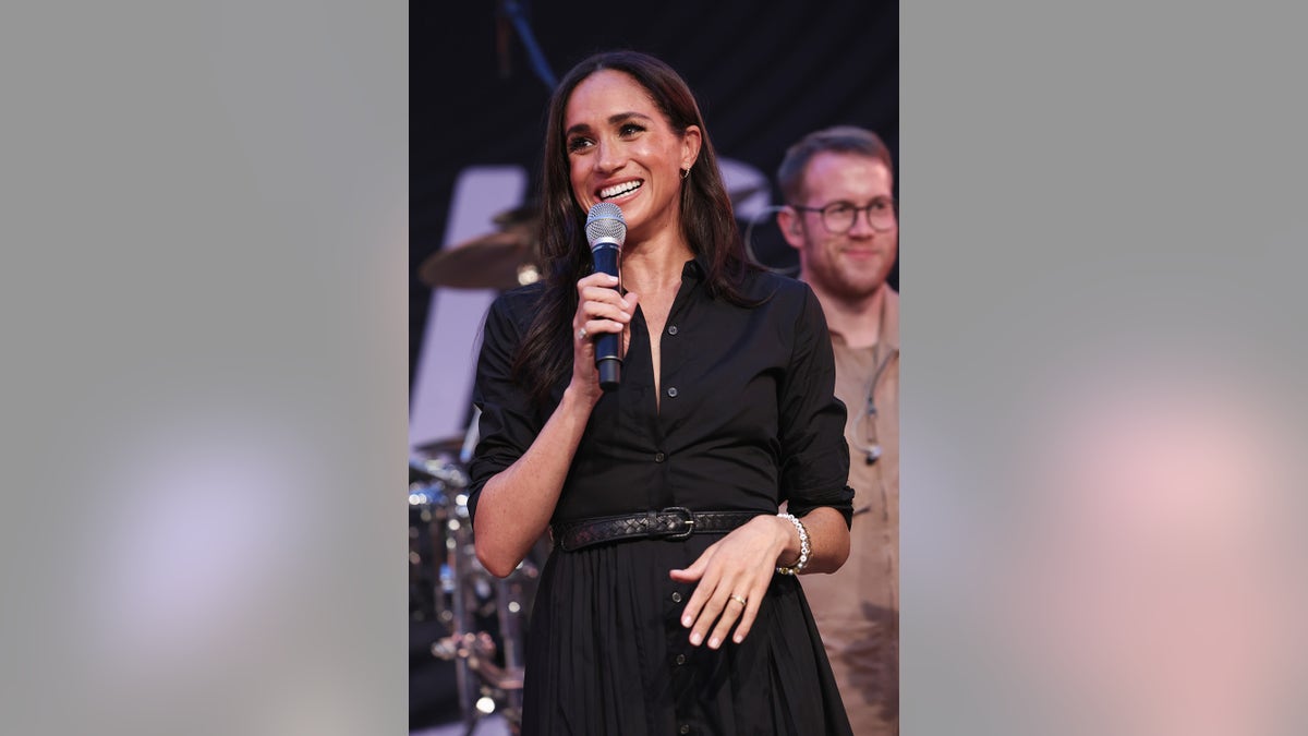 Meghan Markle smiling with a black dress and holding a microphone.