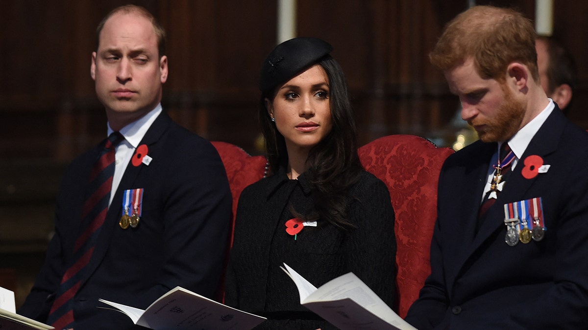 Prince William sitting next to Meghan Markle and Prince Harry in burgundy chairs.