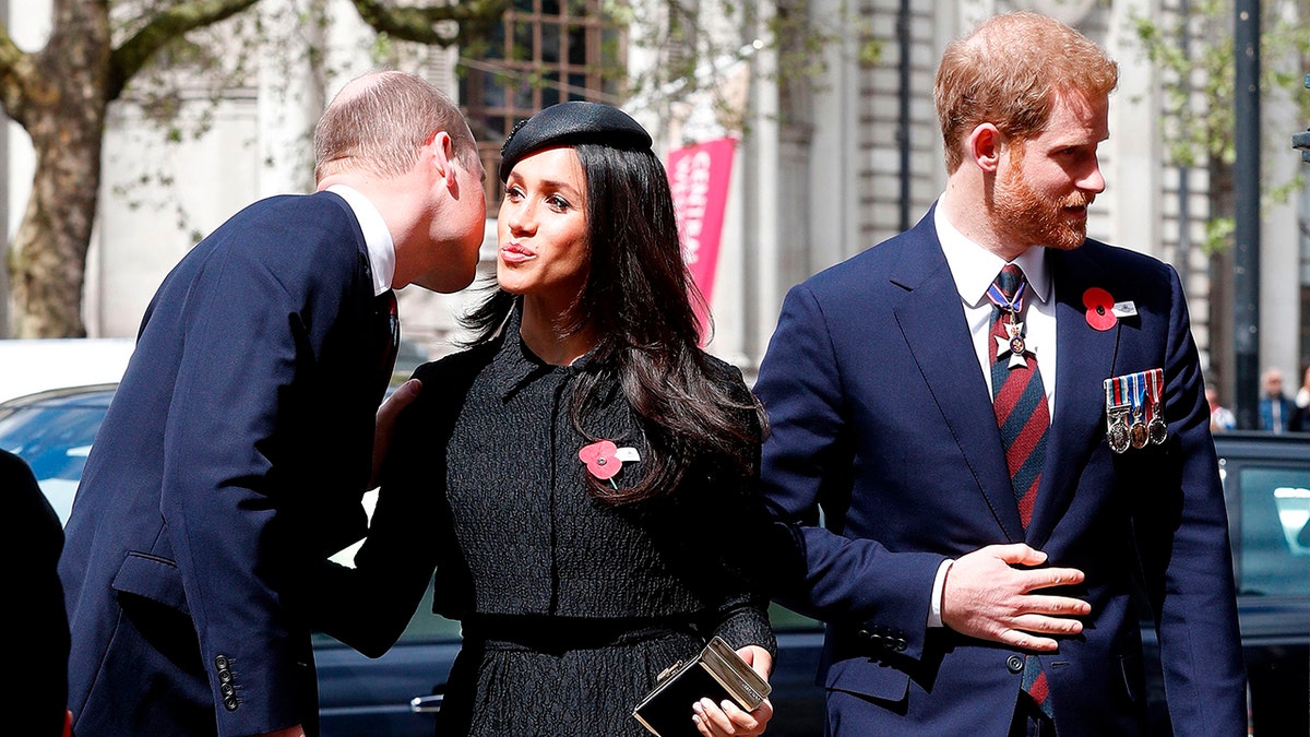 Prince Harry looking away in a suit while Meghan Markle and Prince William formally kiss on the cheek wearing dark suits.