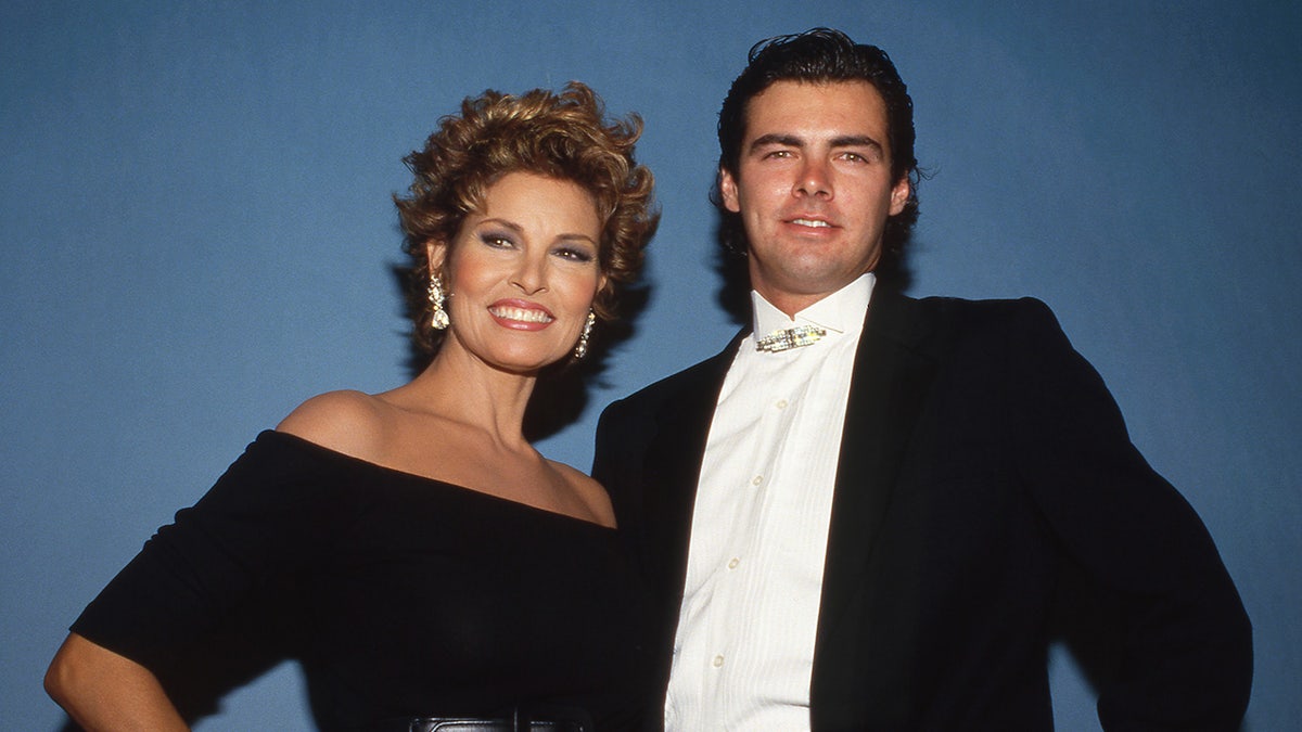 Raquel Welch wearing an off-the-shoulder black dress smiling and standing next to her son Damon in a tux.