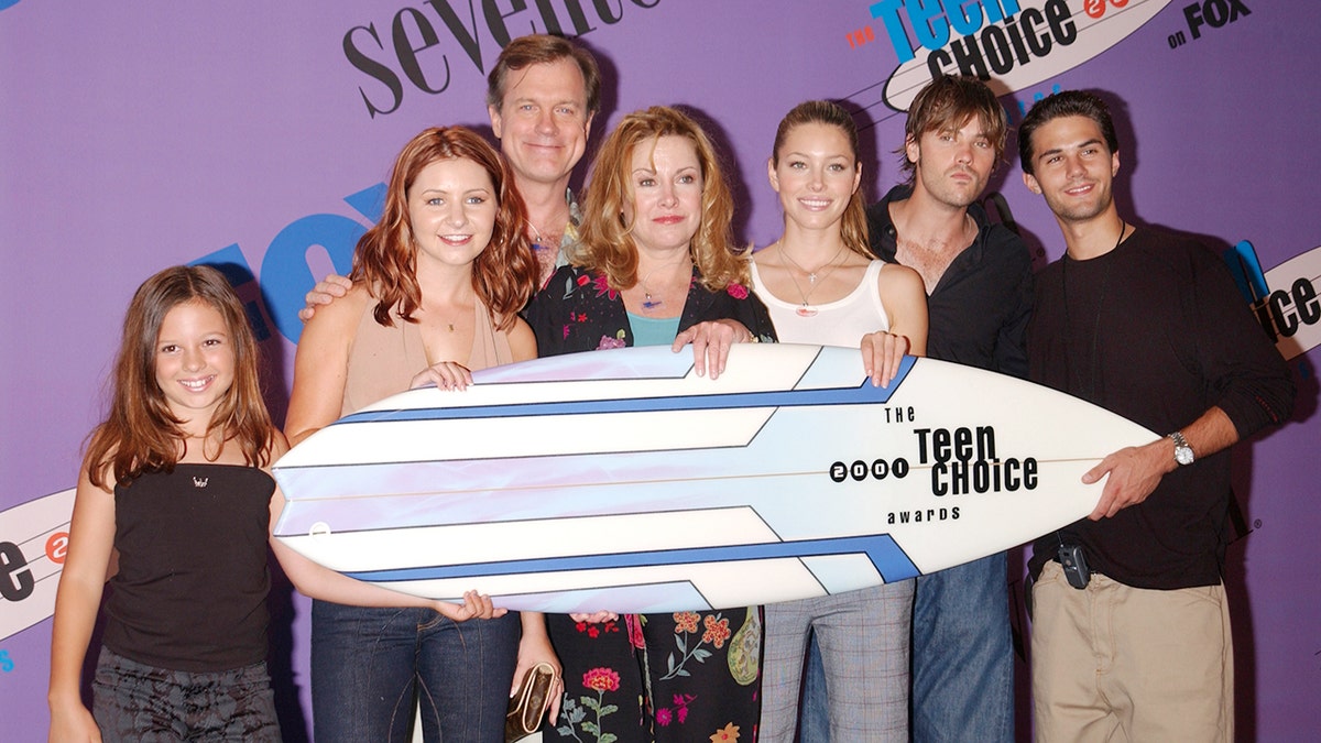 The cast of 7th Heaven smiling together and holding a surfboard.