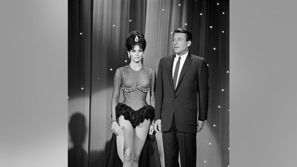 A young Raquel Welch in a sequined showgirl dress on stage next to a man in a suit.