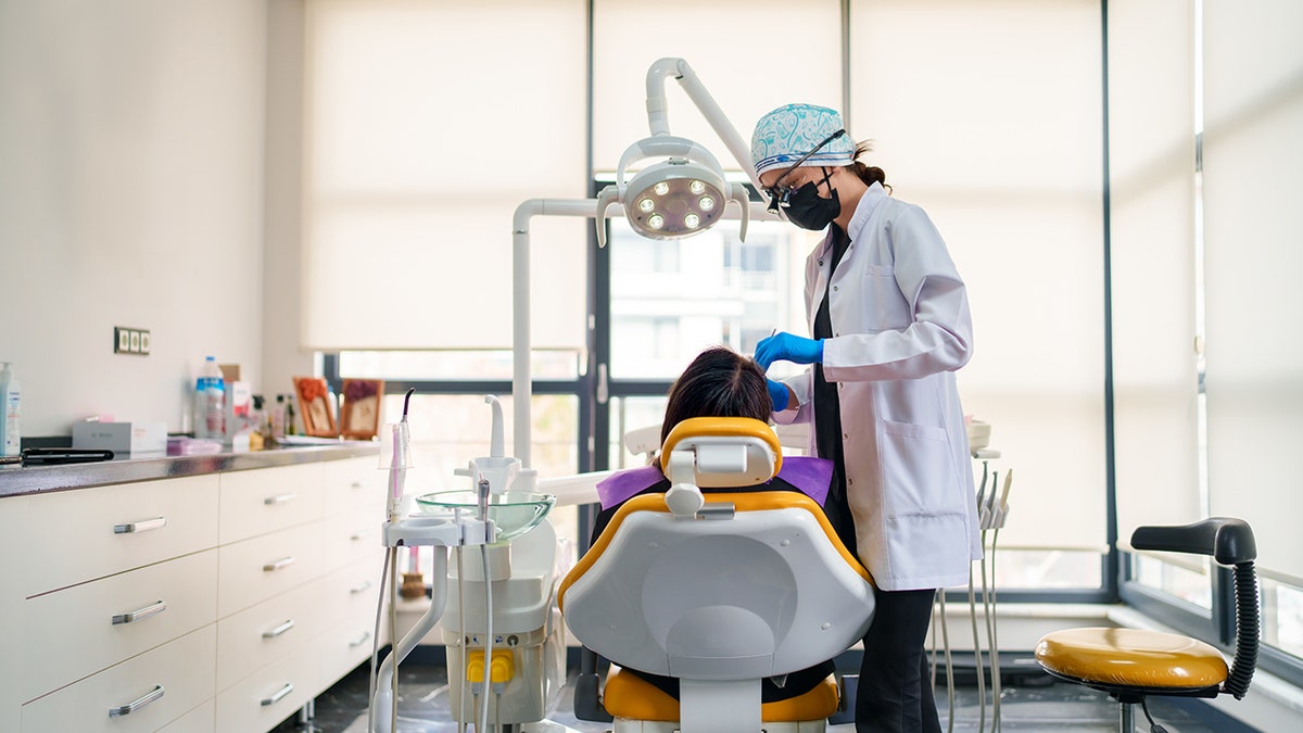 Dentist peers over chair to work on patient's mouth in wide shot of dentist office