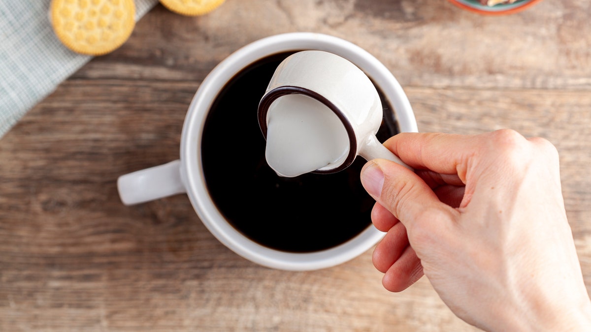 A hand pours creamer into a coffee cup. Cracks are seen in the top left corner.