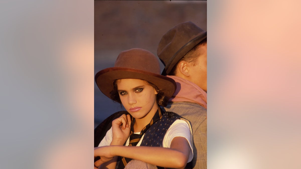 Ione Skye modeling wearing a hat, a white shirt, a dark vest and a black jacket over her shoulder.