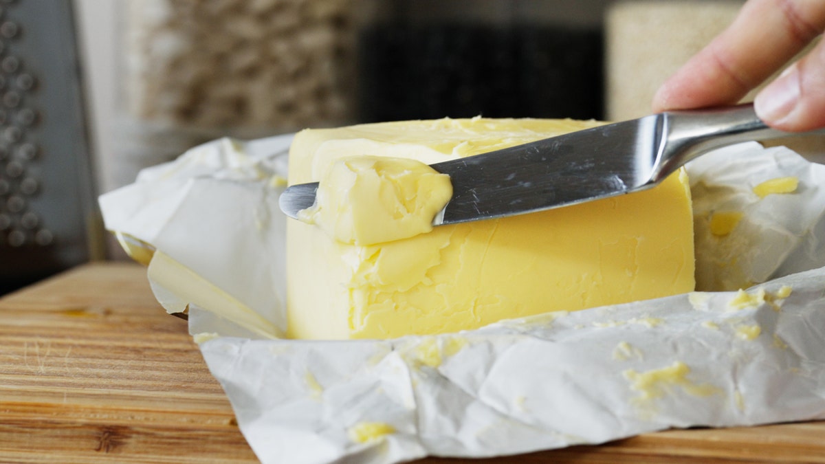 A hand holds a knife cutting into a stick of butter on a block.