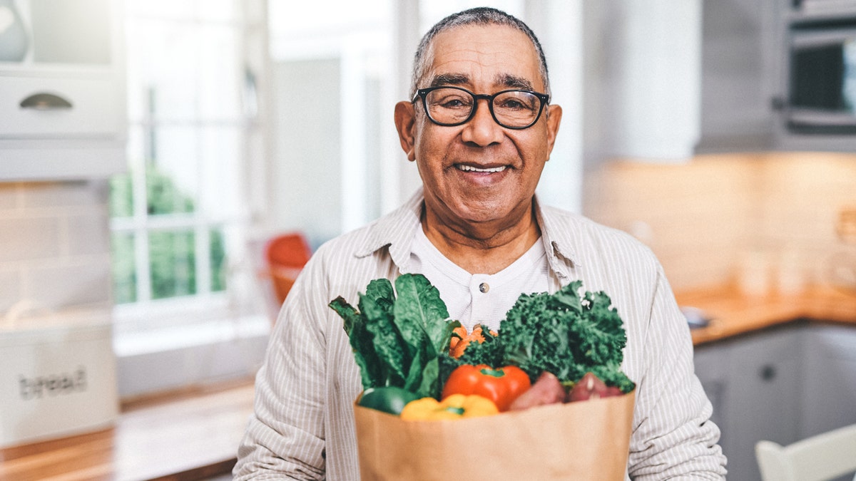 A man wearing glasses holds a grocery bag with fruits and vegetables while standing in a kitchen.