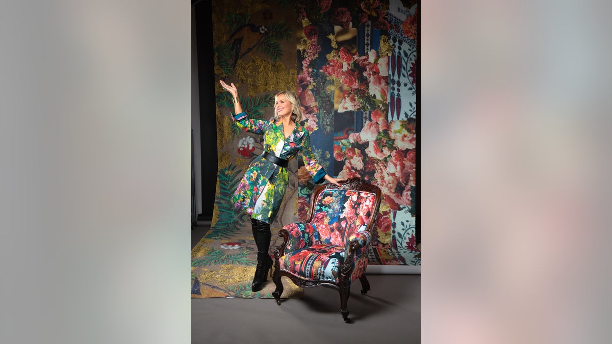 Melora Hardin posing in a collage dress next to a collage printed chair and wallpaper in a dark studio.