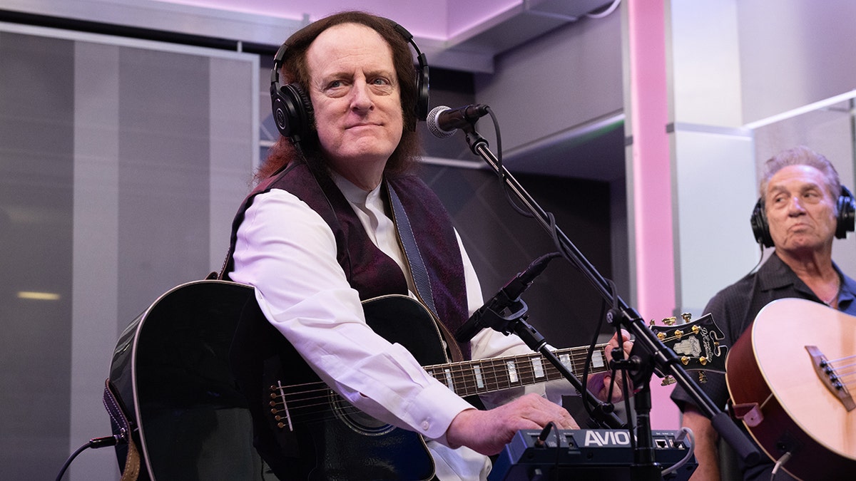 Tommy James sits in a studio with headphones holding a guitar