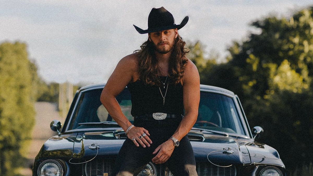 Warren Zeders wears all black and black cowboy hat sitting on a car