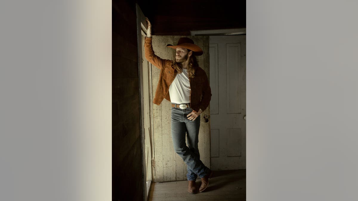 Warren Zeders wears a tan jacket and a cowboy hat, stands at the entrance