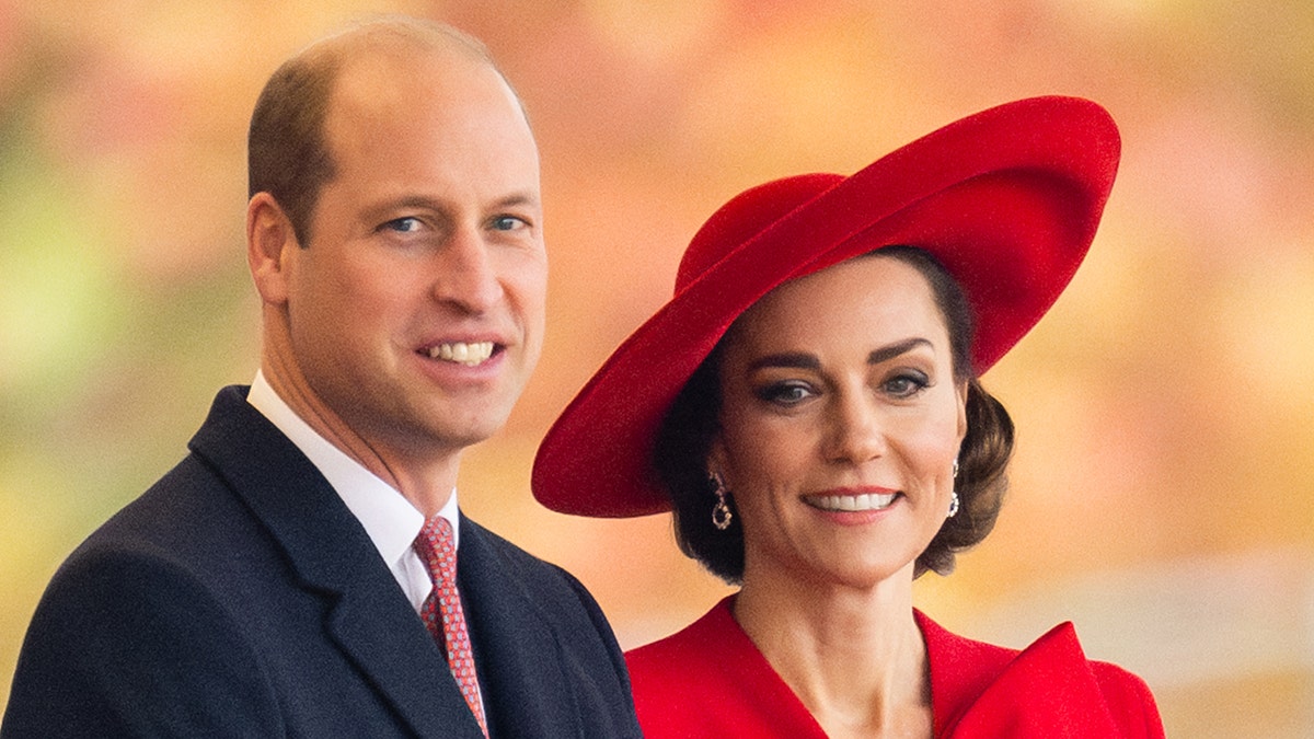 Tampilan dekat Kate Middleton dengan setelan yang berdiri di sebelah Kate Middleton dengan gaun merah dan topi yang serasi.